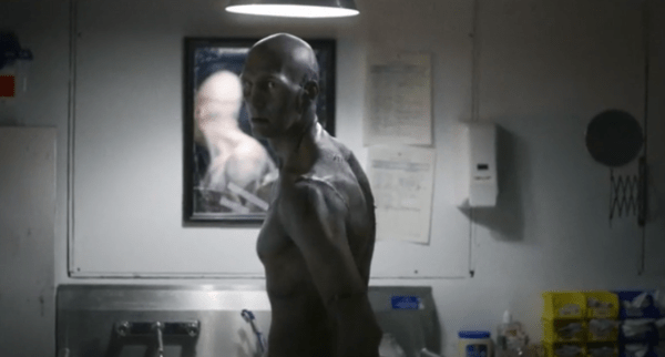 Bald heavily-scarred man with stitches standing in a bathroom in Depraved