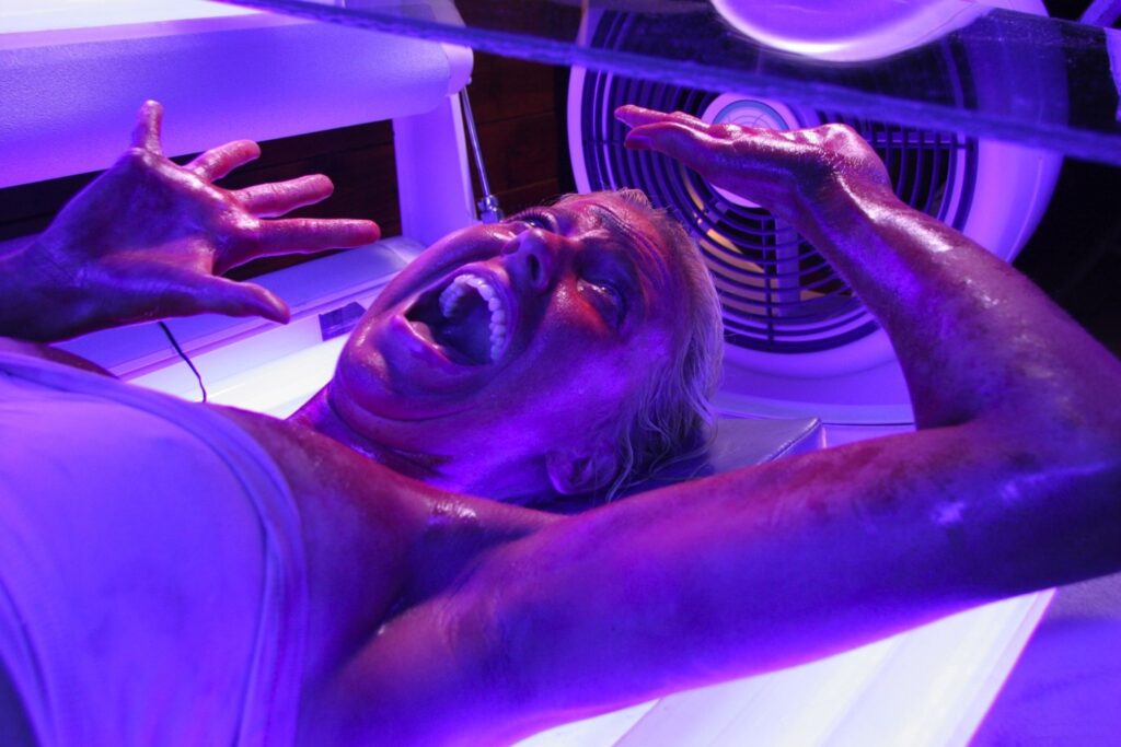 a girl stuck in a tanning booth in Final Destination 3, wide-eyed, open mouth, blue lighting.