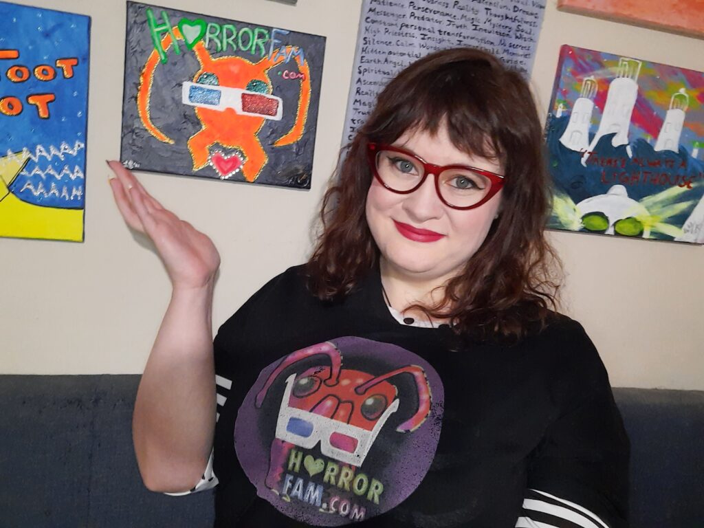 Lauren Spear (a pale woman who's nearly 40 with dark brown wavy hair and mostly-blue eyes) is wearing a HorrorFam.com t-shirt while smiling and gesturing (in a one-handed "ta-da" presenting-type pose) towards an acrylic painting of HorrorFam.com's primary mascot, Zander the ant.